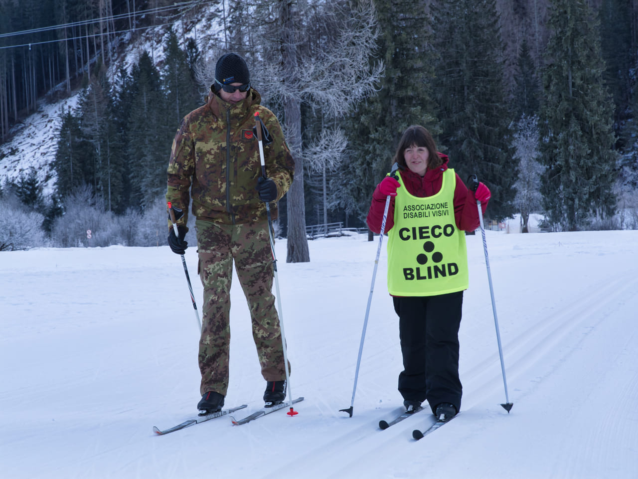 Nell'immagine ci sono due persone che stanno sciando sulla neve. La persona a sinistra indossa un abbigliamento mimetico e un berretto nero, mentre tiene in mano dei bastoncini da sci. La persona a destra indossa un giubbotto giallo fluorescente con la scritta "ASSOCIAZIONE DISABILI VISIVI e tiene anche lei dei bastoncini da sci. Sullo sfondo ci sono alberi innevati e una montagna.