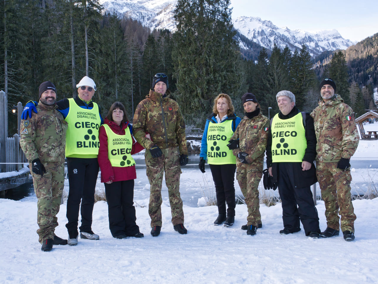 Nell'immagine ci sono otto persone in piedi su un terreno innevato. Sei di loro indossano uniformi mimetiche, mentre le altre due persone indossano abbigliamento invernale. Quattro delle persone indossano pettorine gialle con la scritta "ASSOCIAZIONE DISABILI VISIVI CIECO BLIND" in nero. Sullo sfondo ci sono alberi e montagne innevate. L'atmosfera sembra essere quella di una giornata fredda e invernale.